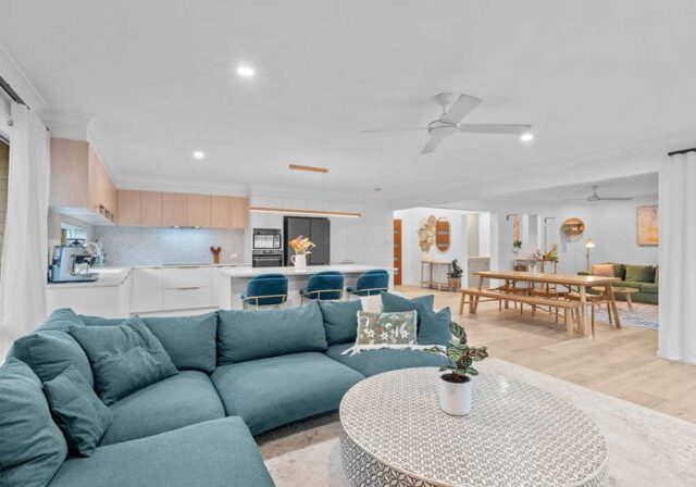 Open-concept living area with a teal sectional couch, round patterned coffee table, and a bright kitchen island with blue bar stools — modern and spacious.