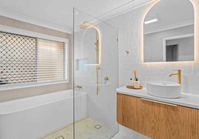 Modern bathroom with glass shower, white tub, and wooden vanity topped with a round white sink and gold fixtures.