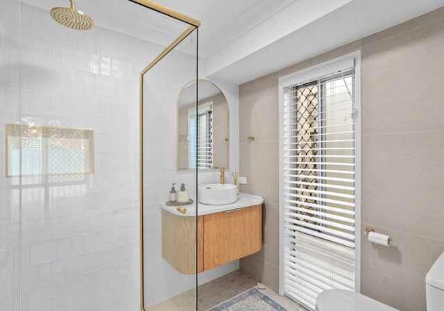 Modern bathroom with a glass shower enclosure, gold fixtures, floating wood vanity, round mirror, and window with blinds.