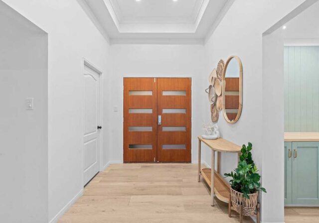 Bright entryway with light wood floors, double wooden doors with horizontal glass panels, and a console table with a round mirror and plant nearby.