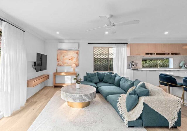 Open-concept living room with a teal sectional sofa, round patterned coffee table, and white curtains.