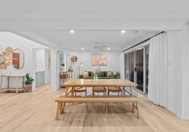 Open-plan dining and living area with a long wooden table and benches, white walls, light wood floors, and sliding glass doors on the right.