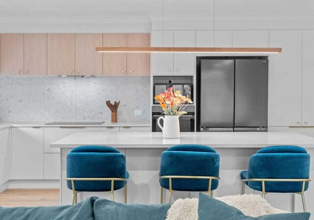 Modern white kitchen with a large island, blue velvet bar stools, and a stainless steel fridge.