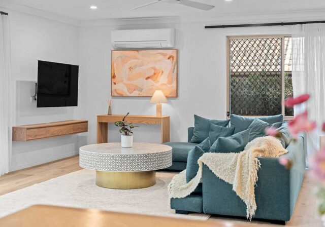 Modern living room with a teal sofa, round patterned ottoman, cream rug, and a wooden console under a wall-mounted TV.
