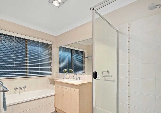 Bathroom with a tub beneath two large windows, a light wood vanity, and a glass shower enclosure.
