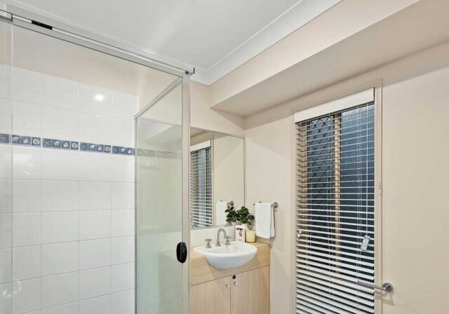 Modern bathroom with a glass shower enclosure, white tiled walls, and a small wooden vanity with a sink and mirror across from a window with blinds.
