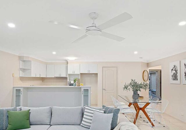 Bright open-plan living area with a white kitchen, glass dining table and white wire chairs, and a large ceiling fan overhead.