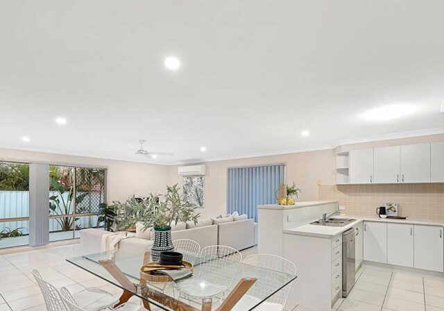 Open-plan living and kitchen area with white cabinetry, glass dining table, white wire chairs, indoor plants, and sliding glass doors to a patio/outdoor space.