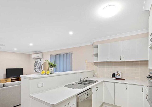 Bright open-plan kitchen with white cabinets, beige tiled backsplash, double sink, and dishwasher, opening to a living area with a TV.