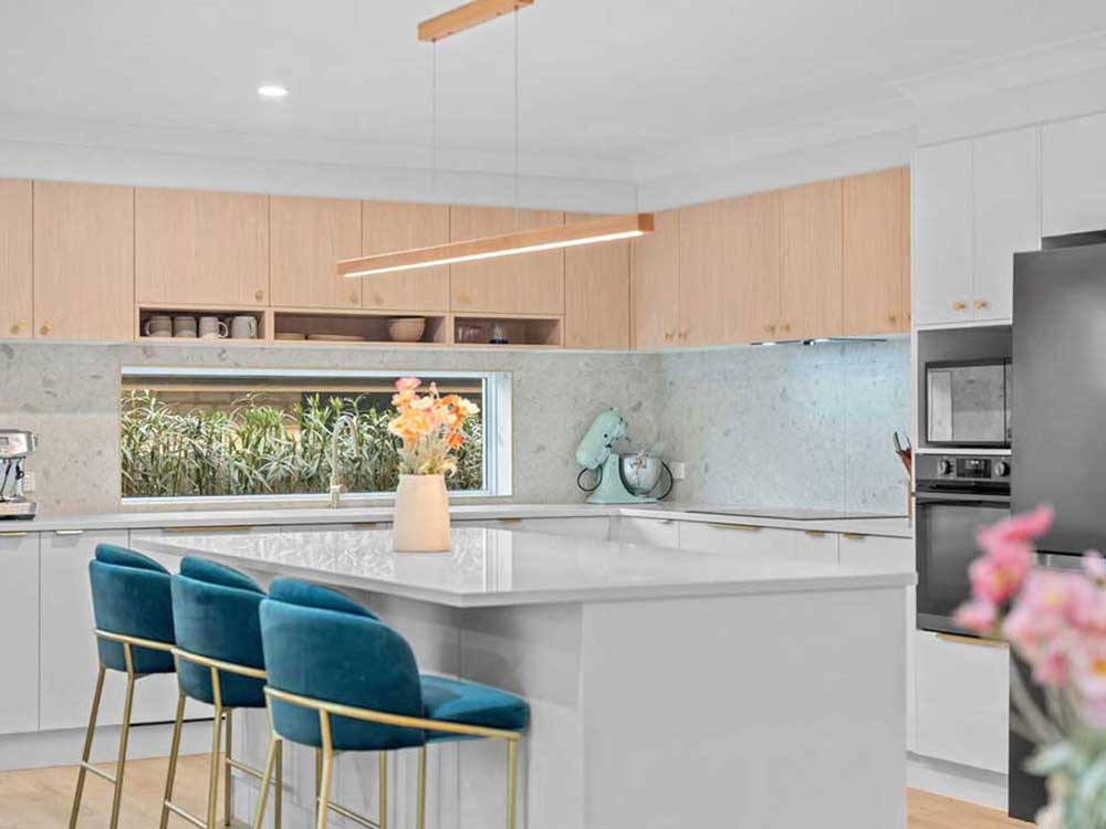 Modern kitchen with a white island, teal bar stools, light wood cabinets, and a vase of flowers on the island.
